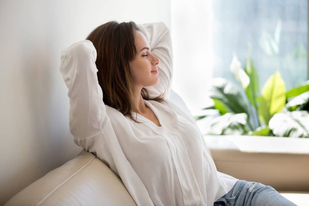 une femme se relaxe détendue sur un canapé, les mains derrière la tête et les yeux fermés