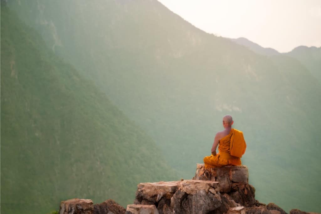 un moine bouddhiste en tenue orange et crane rasé est assis en méditation sur des rochers. devant lui, des montagnes imposantes le surplombent.