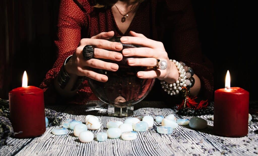mains de médium portant des bracelets dans un décor sombre posées sur une boule de cristal avec deux bougies, plusieurs pierres blanches et bleues posées sur la table