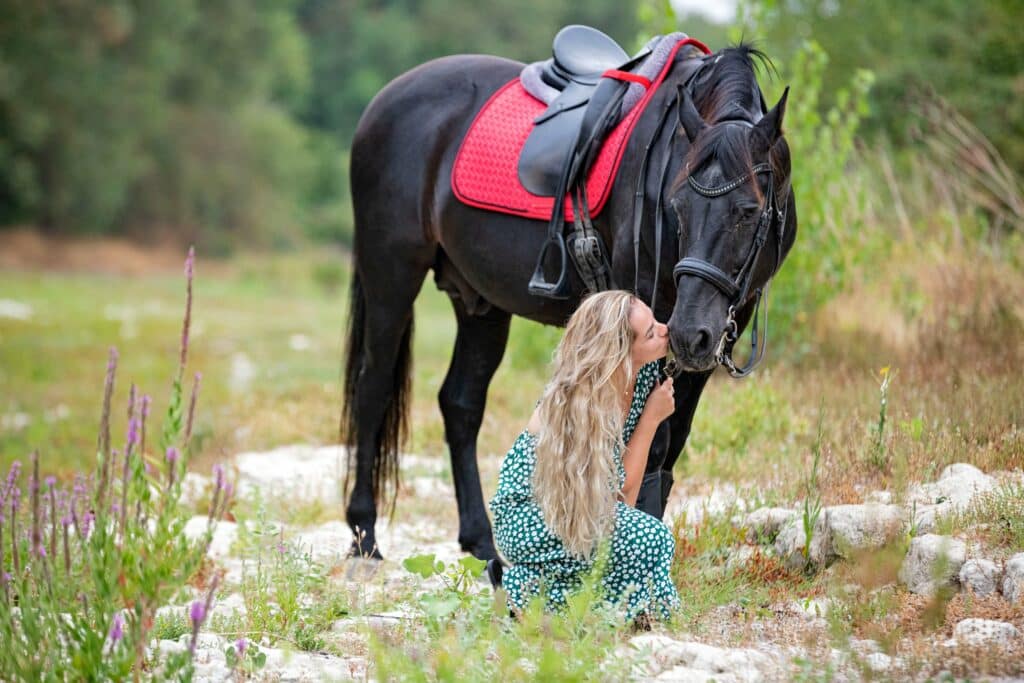 une femme blonde est accroupie et embrasse un cheval noir sur le museau. le cheval porte une selle. ils sont dans la nature.
