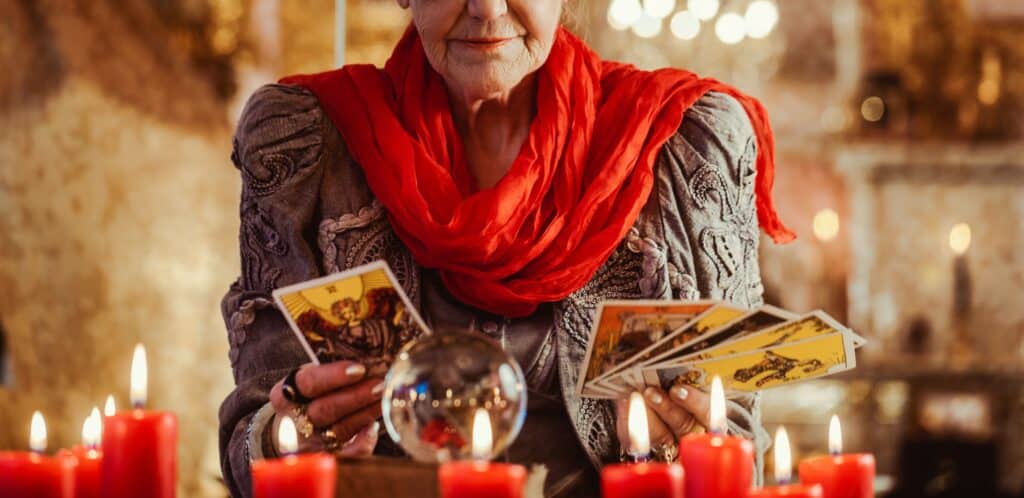 une voyante tient des cartes devant des bougies et une boule de cristal pour une séance de voyance. Elle porte un foulard orange et un haut kaki. 