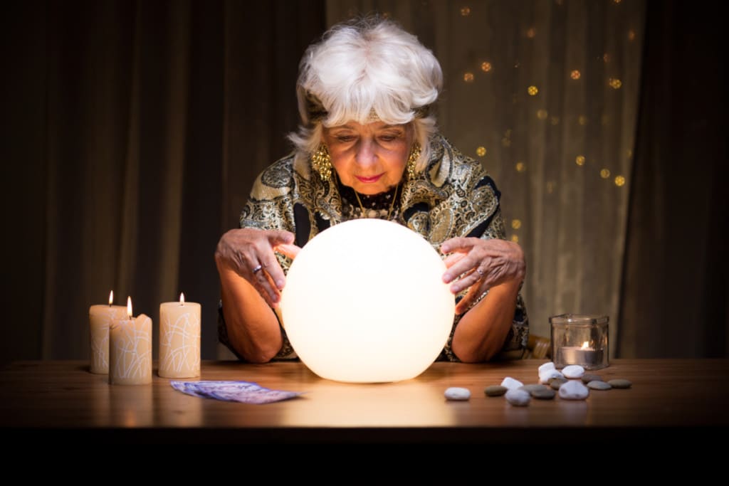 une femme âge voyante tient dans ses mains une grande boule de cristal illuminée. des bougies allumées, pierres et cartes sont posées sur sa table.