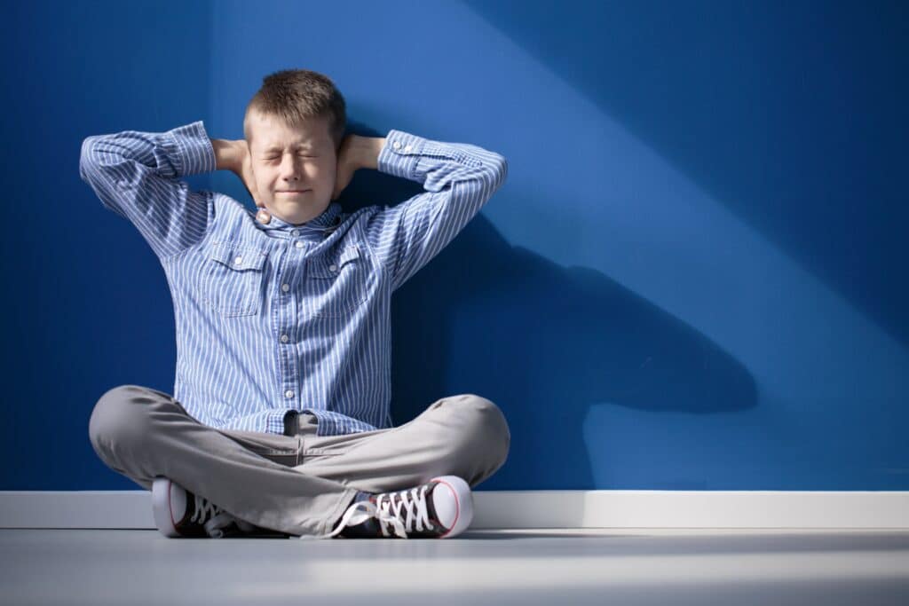 Un garçon avec une hypersensibilité auditive, assis sur le sol contre un mur bleu, se couvrant les oreilles avec ses mains et fermant ses yeux.