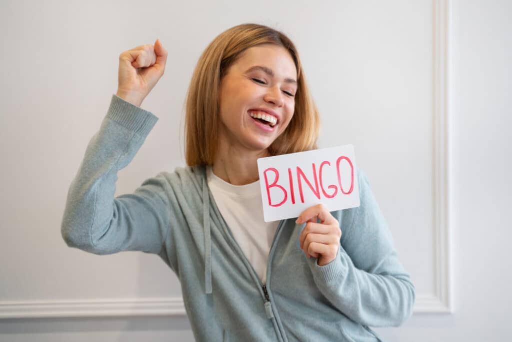 Une femme souriant, fermant les yeux et levant le poing en signe de victoire, tenant un papier où est écrit le mot "Bingo" pour exprimer la chance.